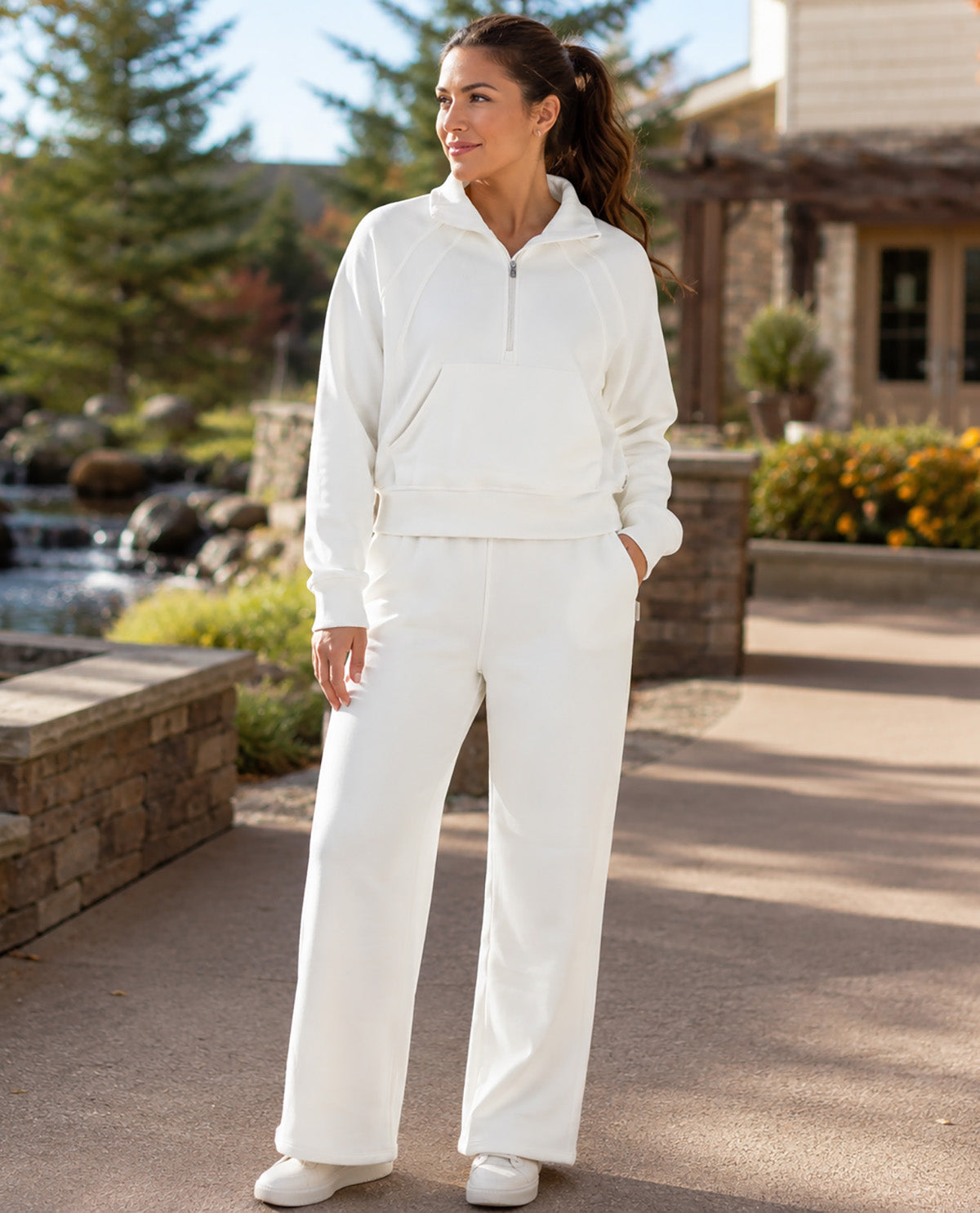 A woman wearing a matching white lounge set consisting of a half-zip hoodie and wide-leg pants, standing outdoors on a paved walkway in front of a resort-style building.