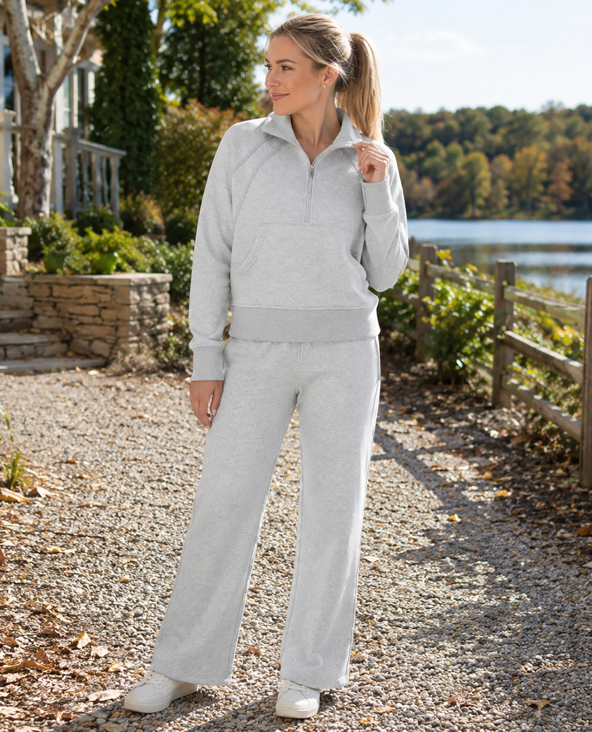 A woman wearing a light gray matching lounge set consisting of a quarter-zip long-sleeve top and wide-leg sweatpants, standing on a gravel path outdoors near a lake.