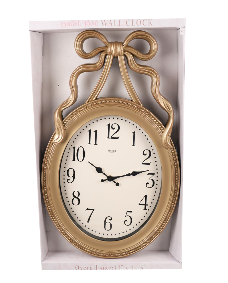 A gold-framed wall clock with a decorative bow-shaped top and a white clock face, shown inside its white retail box.