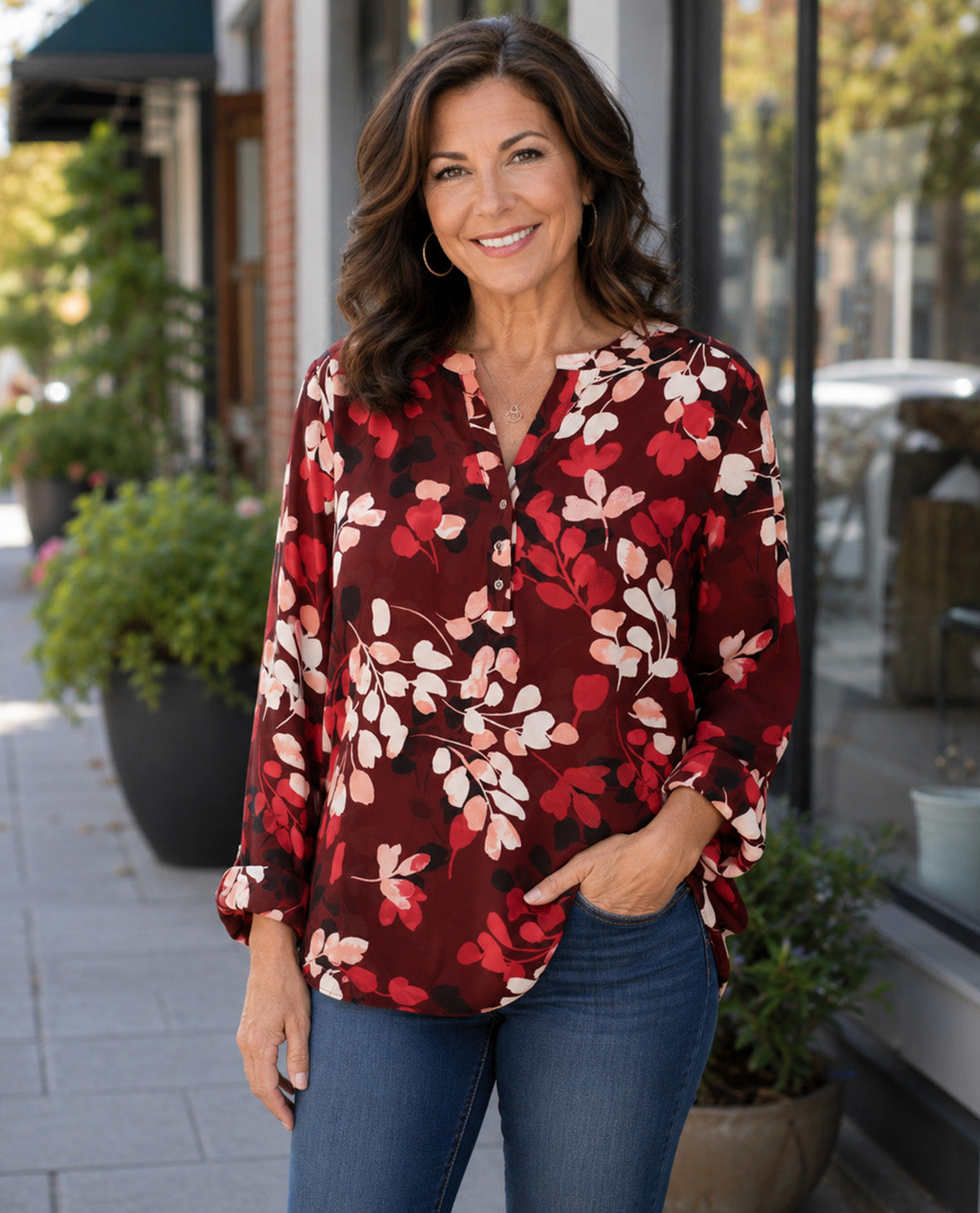 A woman wearing a long-sleeve button-up blouse with a red, pink, and white floral pattern, paired with blue jeans, standing outdoors on a sidewalk in front of a storefront.