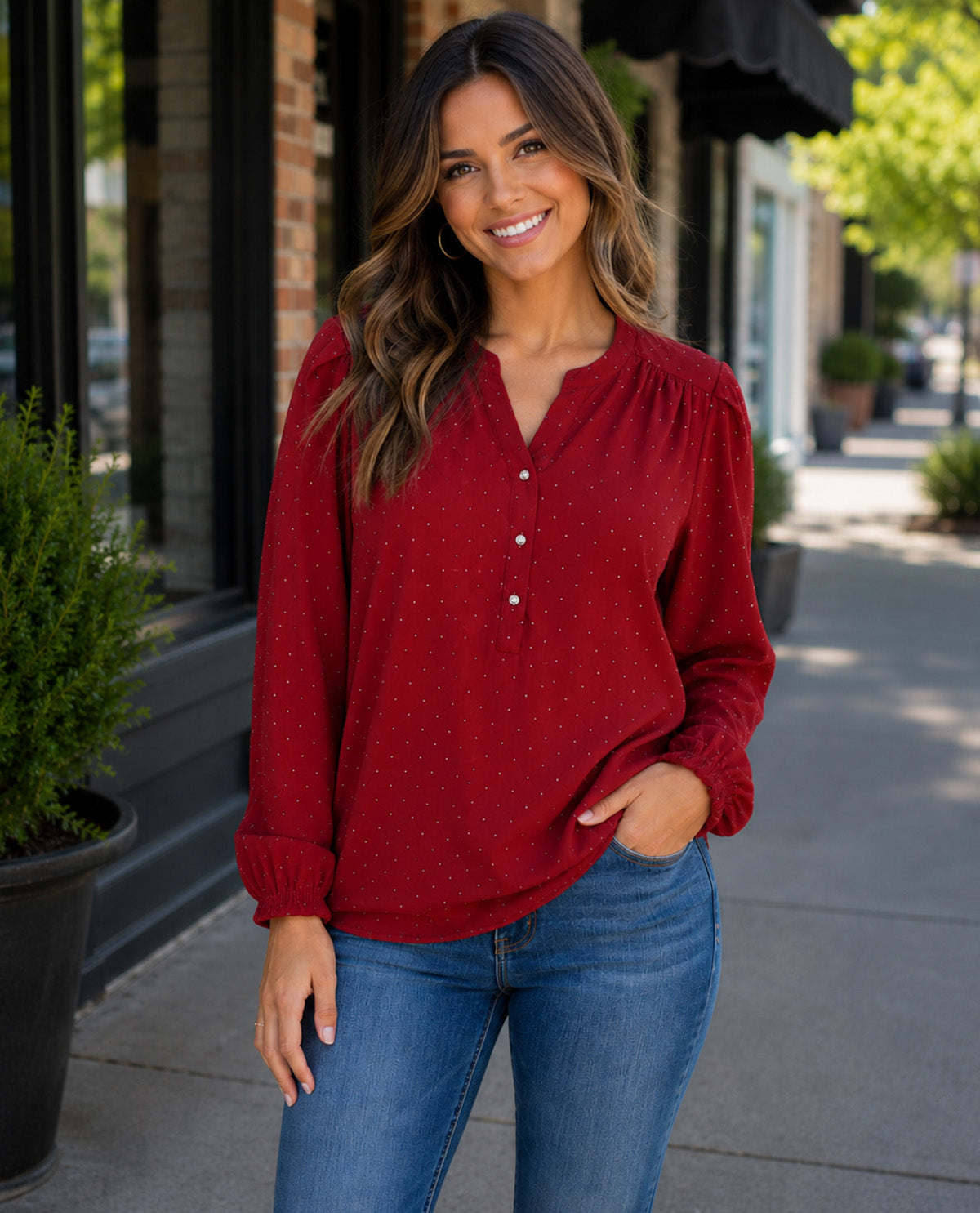 A woman wearing a red long-sleeve blouse with a small white polka dot pattern and button placket, paired with blue jeans, standing on a city sidewalk.