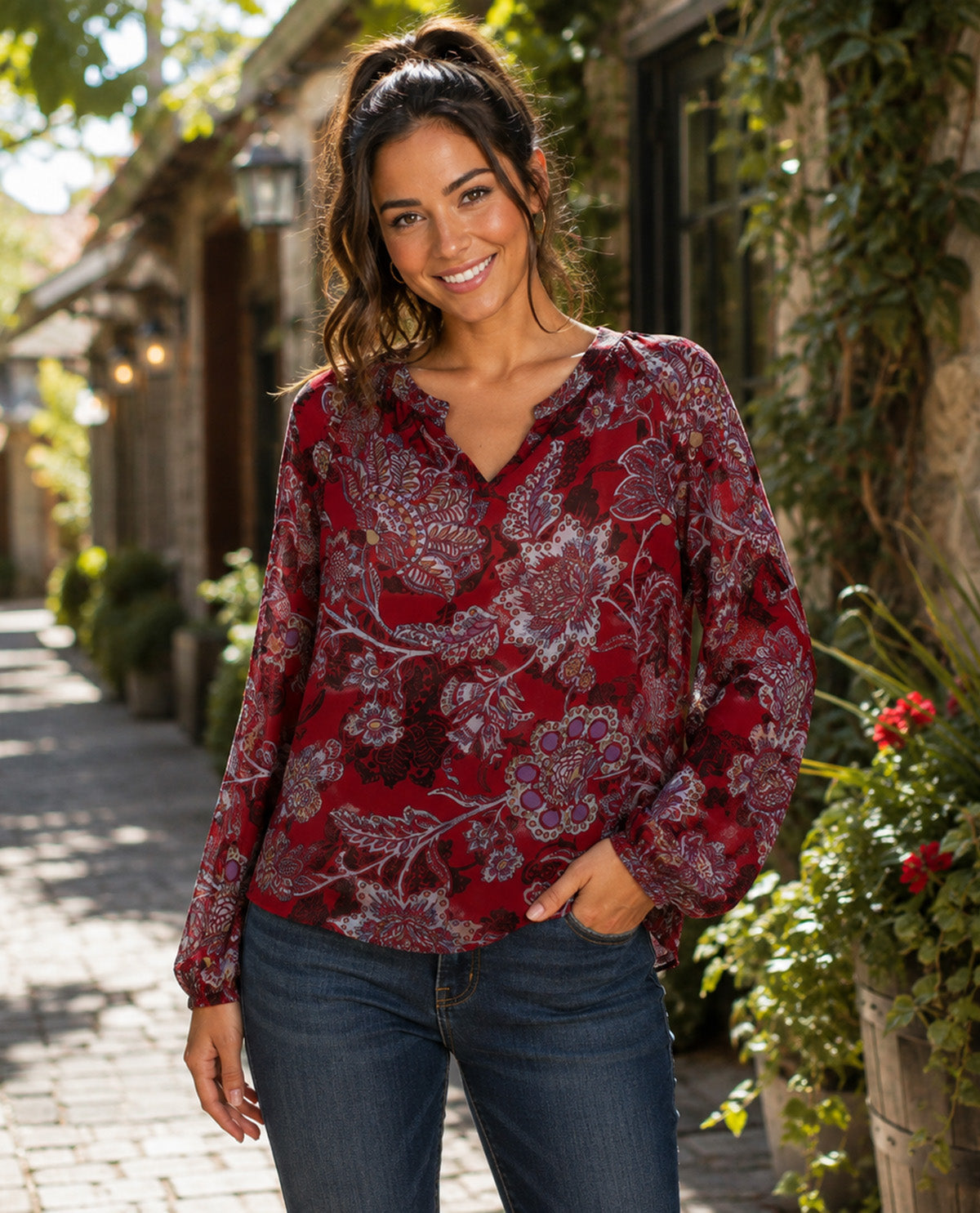 A woman wearing a long-sleeve red floral blouse and blue jeans, standing in a sunlit outdoor alley with stone walls and greenery.