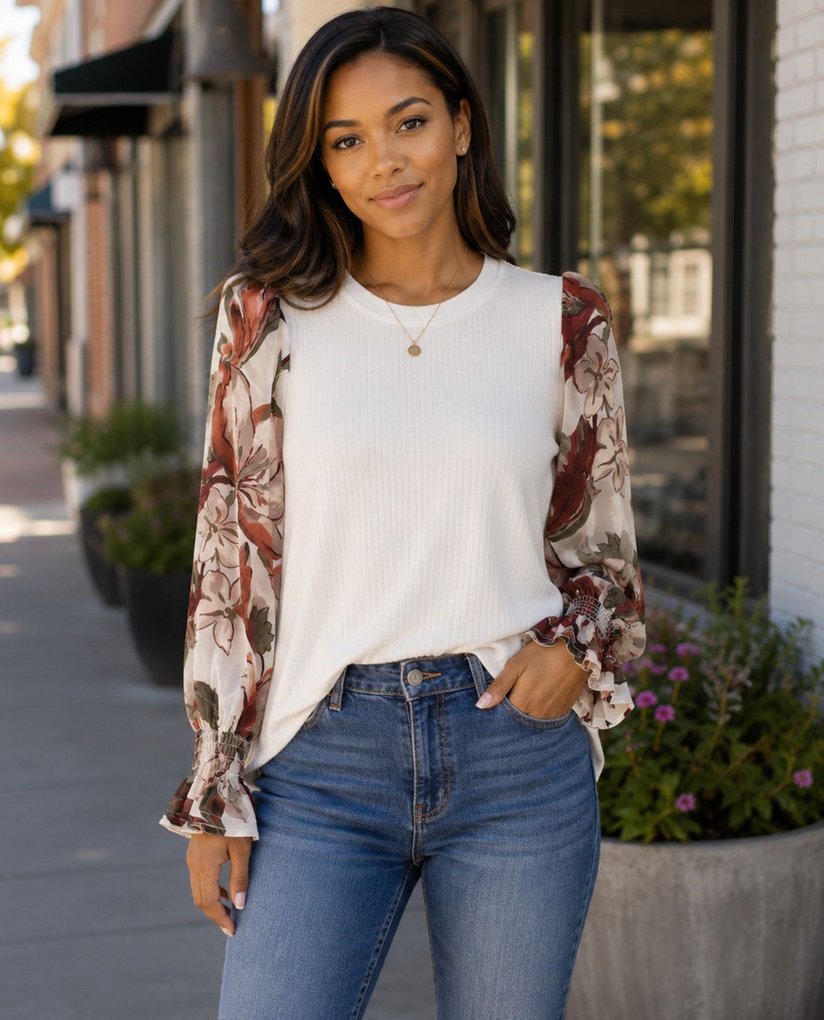 A woman modeling a white ribbed long-sleeve top with sheer floral bell sleeves, paired with blue jeans, standing on a city sidewalk.