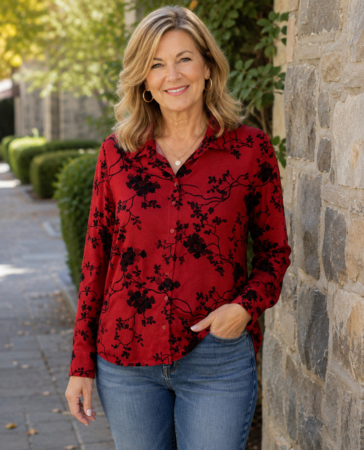 A woman wearing a red long-sleeve button-up blouse with a black floral branch pattern, paired with blue jeans, standing outdoors next to a stone wall.