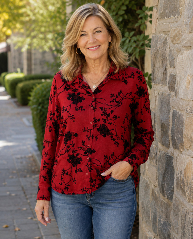 A woman wearing a red long-sleeve button-up blouse with a black floral branch pattern, paired with blue jeans, standing outdoors next to a stone wall.