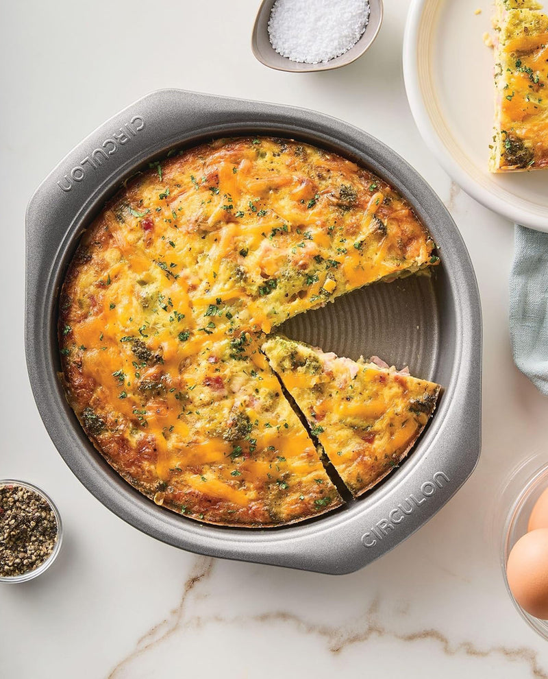 A baked breakfast pie or frittata in a round gray nonstick pan, shown from above on a light marble surface with a slice cut out. Small bowls of salt, pepper, and broccoli are arranged around it, and part of a plate with another slice is visible at the top right.
