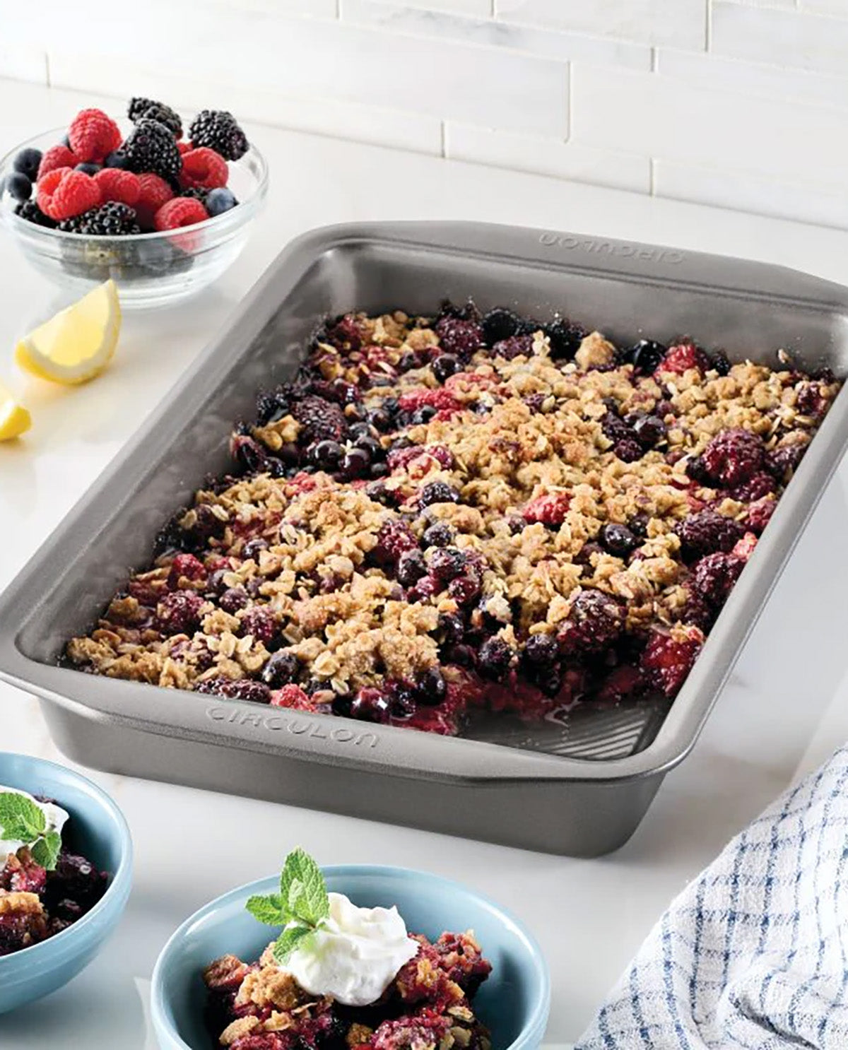 A rectangular stainless steel baking pan filled with a berry crisp dessert, styled on a white countertop with bowls of fresh berries, whipped cream, and a lemon wedge.