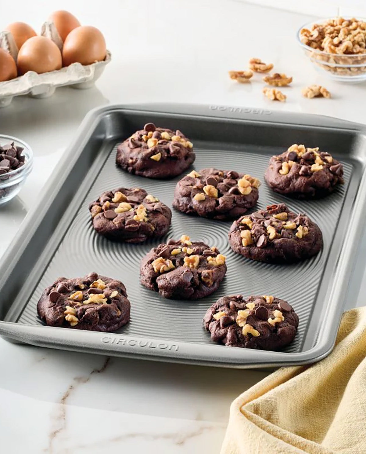 A round gray silicone baking pan with embossed concentric circles and the brand name 'CICULON' on the bottom rim, holding seven chocolate chip cookies topped with chopped walnuts. The pan is styled on a white marble countertop with baking ingredients like eggs, walnuts, and flour in the background.