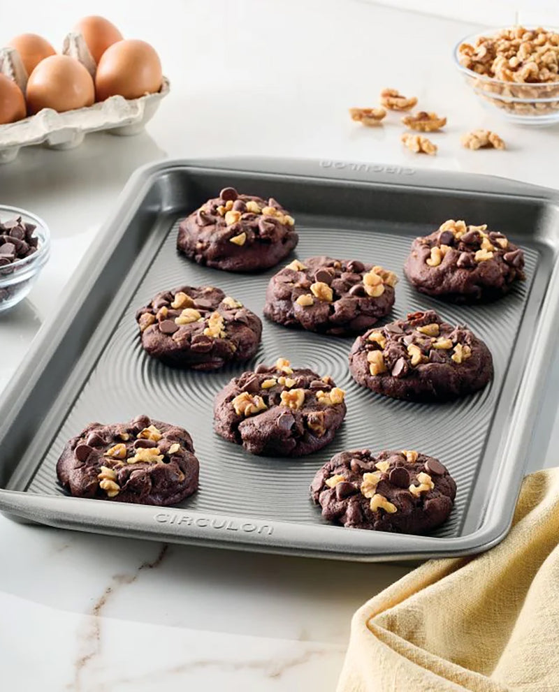 A round gray silicone baking pan with embossed concentric circles and the brand name 'CICULON' on the bottom rim, holding seven chocolate chip cookies topped with chopped walnuts. The pan is styled on a white marble countertop with baking ingredients like eggs, walnuts, and flour in the background.