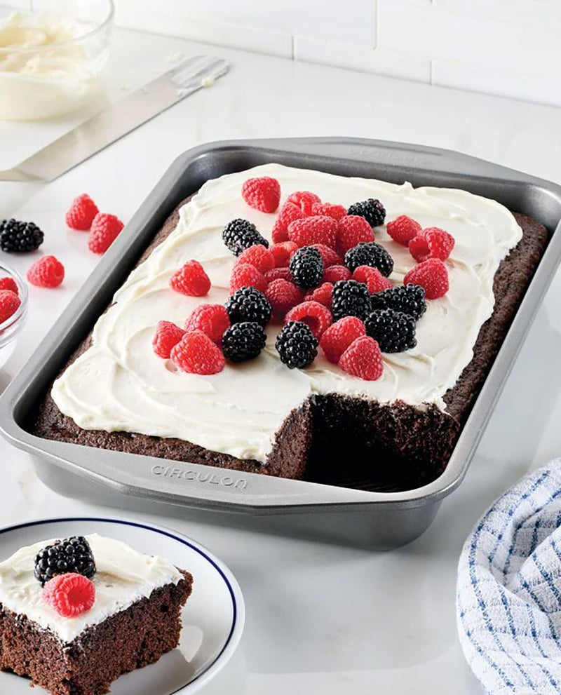A square baking pan containing a chocolate cake topped with white frosting and fresh raspberries and blackberries, with a slice served on a plate in the foreground on a light countertop.