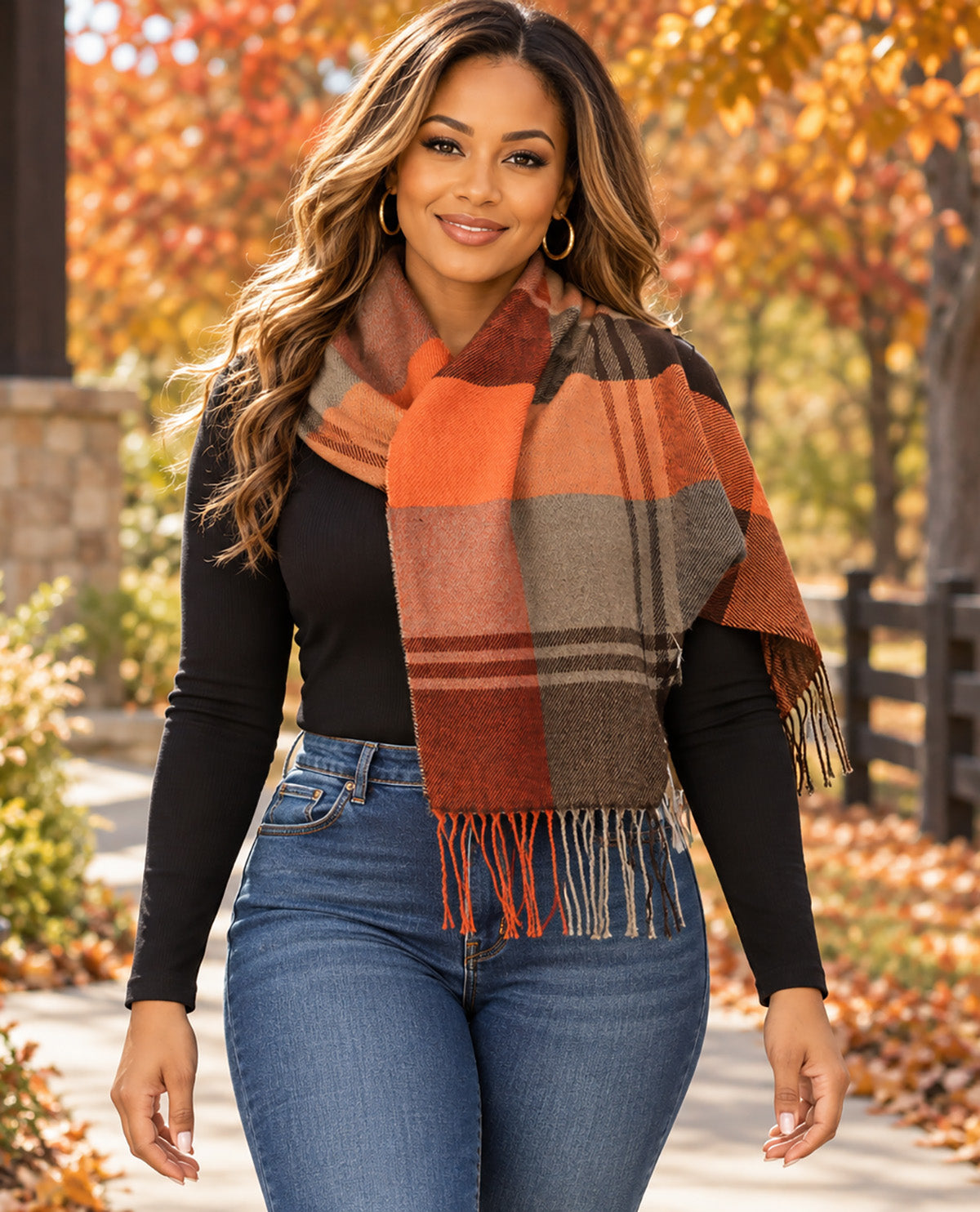 A woman wearing a large plaid scarf in autumn colors, including rust orange, brown, and gray, styled with a black long-sleeve top and blue jeans in an outdoor park setting with fall foliage.
