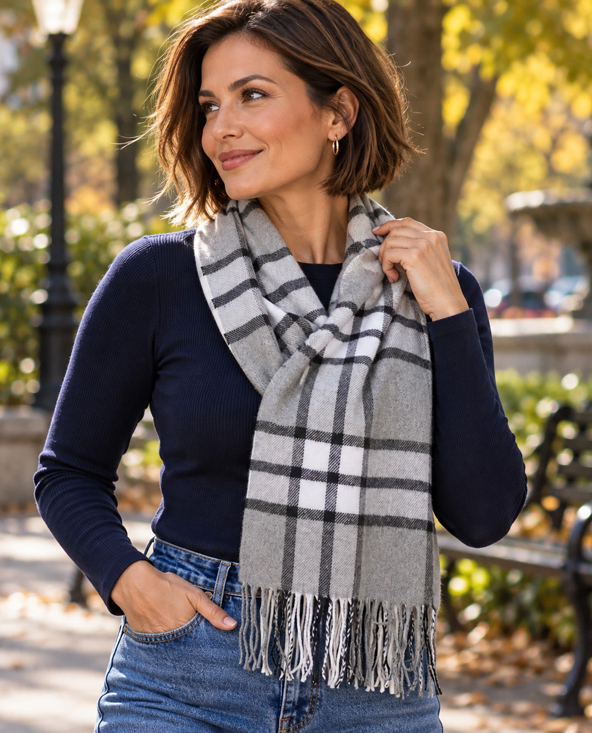 A woman wearing a gray, white, and black plaid fringed scarf over a navy blue long-sleeve top and blue jeans, standing in a sunny park with trees and benches in the background.