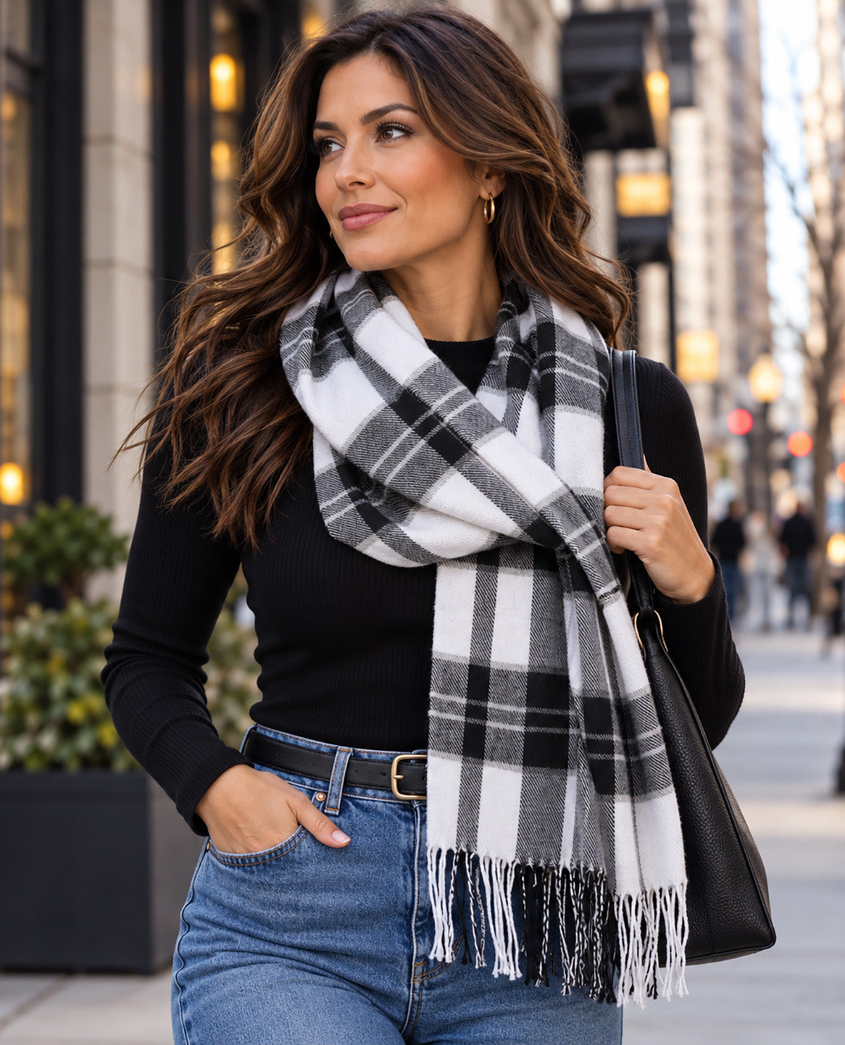 A woman wearing a black long-sleeve top, blue jeans, and a large black, white, and gray plaid fringed scarf, carrying a black shoulder bag on a city street.