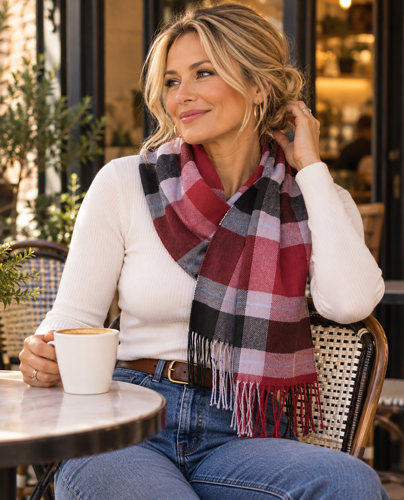 A woman sitting at an outdoor cafe table wearing a red, black, and white plaid scarf, a white ribbed sweater, and blue jeans, holding a white mug.