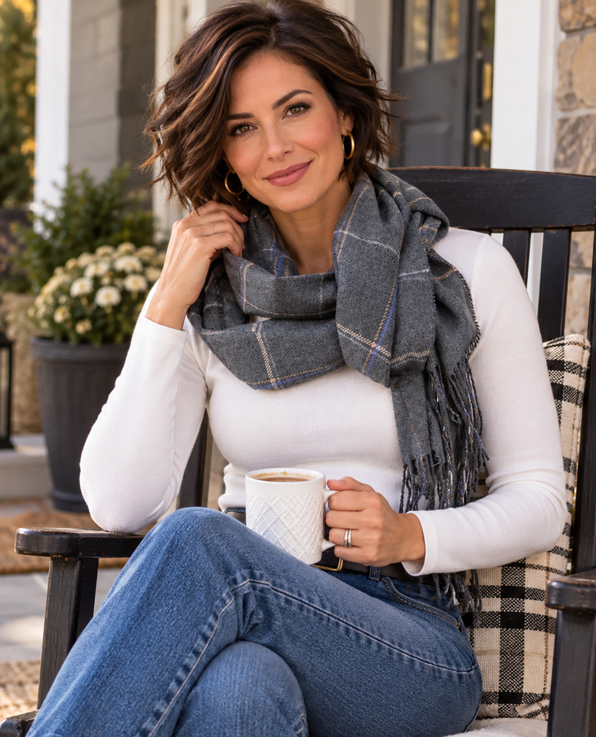 A woman sitting on a porch chair wearing a gray plaid scarf, a white long-sleeve top, and blue jeans, holding a white mug.