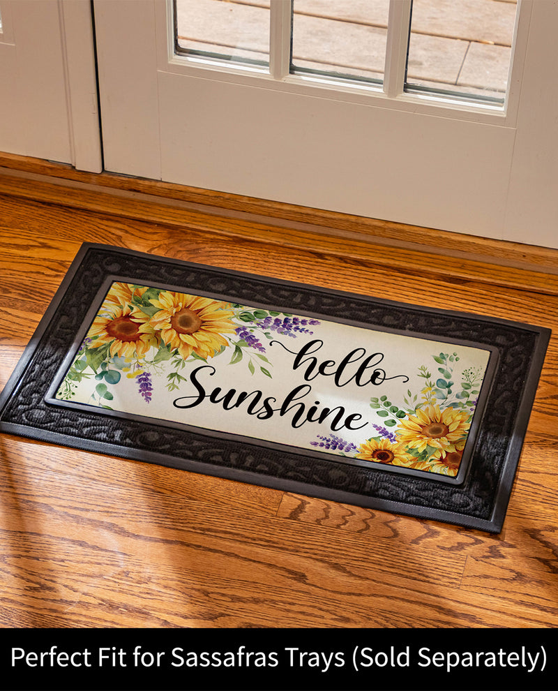 A rectangular decorative doormat with a black ornate border and the phrase “hello Sunshine” in black script, decorated with yellow sunflowers, green leaves, and purple flowers. The mat is shown on a wooden floor in front of a white door, with a text banner at the bottom stating “Perfect Fit for Sassafras Trays (Sold Separately)”.