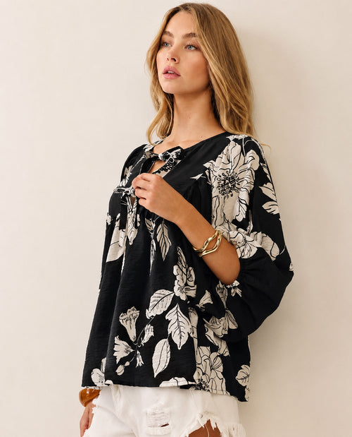 A woman standing wearing a black and white floral long-sleeve blouse with white shorts