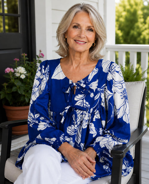 A smiling older woman seated in a porch chair wearing a blue and white floral long-sleeve blouse with white pants, photographed outdoors on a home porch.