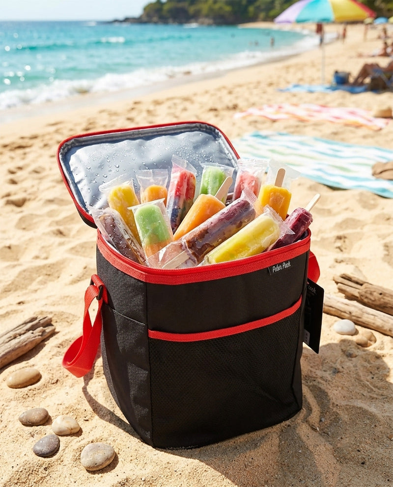 A black cooler bag with red trim sits open on a sandy beach, filled with colorful popsicles. The background shows the blue ocean, shoreline, and beach umbrellas.