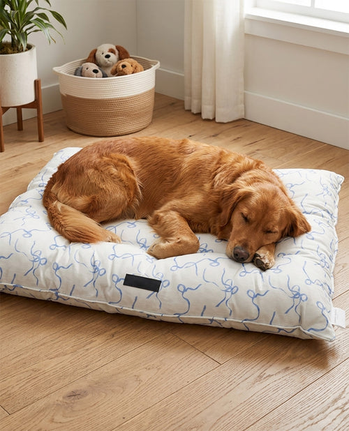A golden retriever dog sleeping comfortably on a white rectangular dog bed with a blue abstract line pattern, placed on a light wood floor in a bright room.