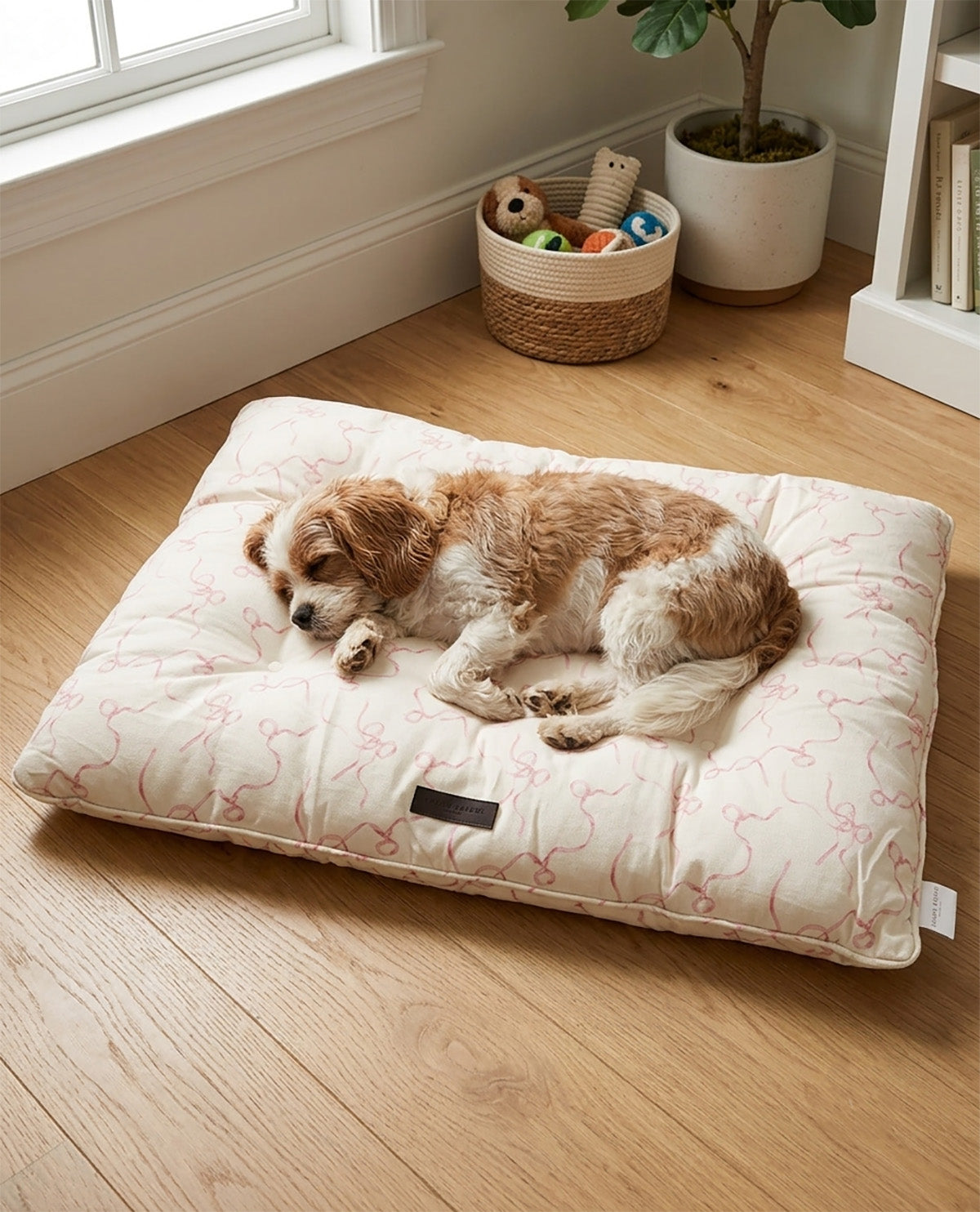 A small brown and white dog rests on a large cream-colored dog bed with a pink bone pattern, placed on a light wood floor in a bright room.
