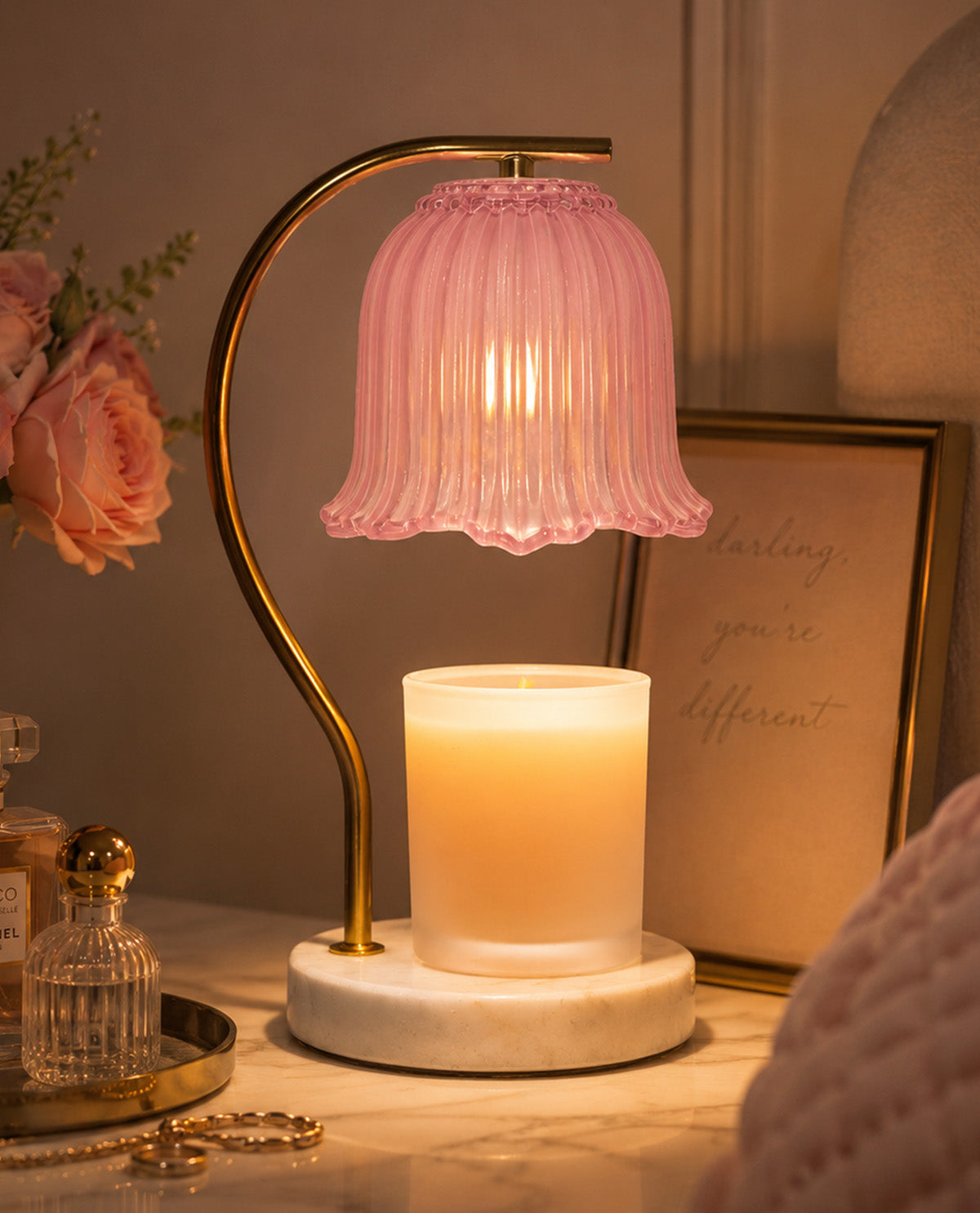 Table lamp with pink glass shade and candle on a marble base, surrounded by decorative items.