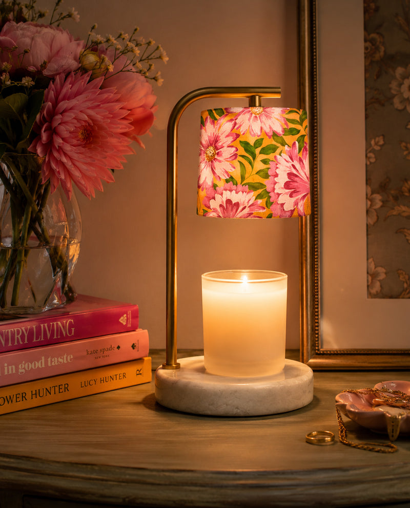 Candle in a holder on a marble surface with a floral lampshade, books, and flowers in the background.