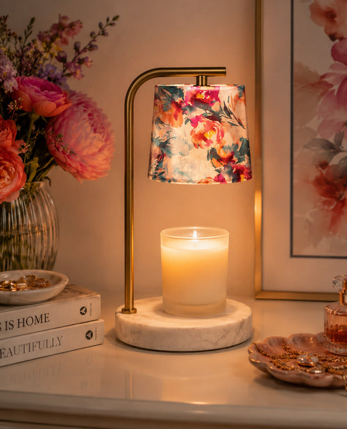 Decorative table lamp with floral shade on a marble base, next to a candle and books, with a floral painting in the background.