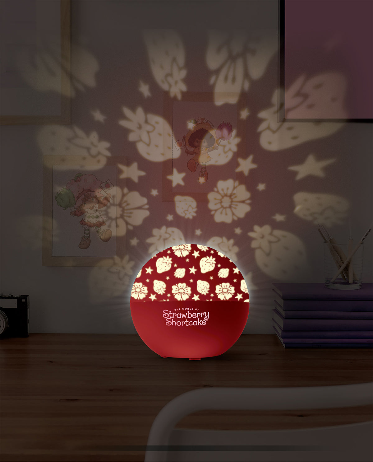 A red spherical Strawberry Shortcake night light sits on a wooden table, casting floral and fairy patterns onto a dark wall in a bedroom setting.