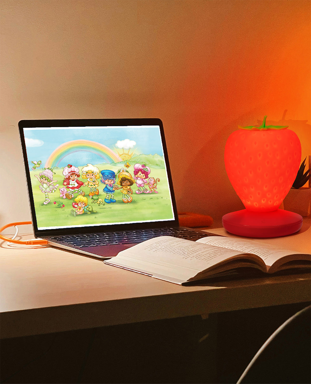 A strawberry-shaped red LED lamp glowing on a desk next to an open laptop showing a cartoon scene and an open book, with warm orange ambient light reflecting on the wall.