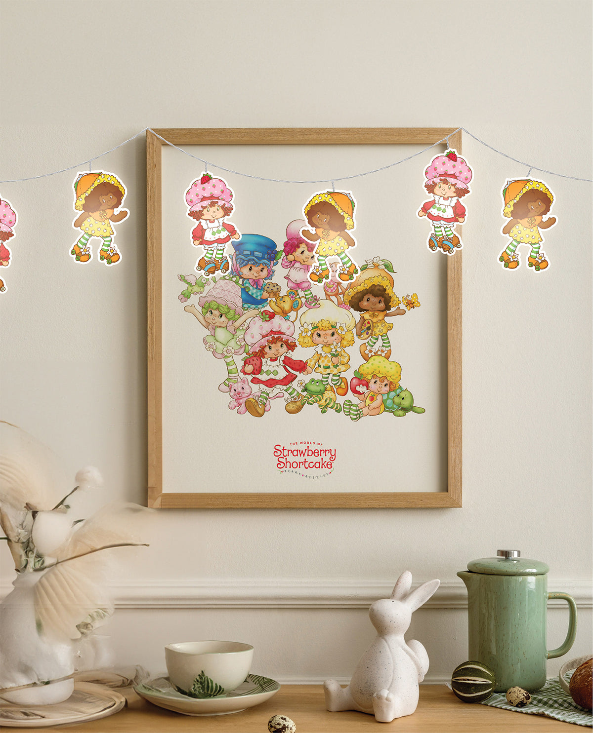 A framed Strawberry Shortcake character print hanging on a cream wall above a tabletop with decorative props, including a white rabbit figurine, a green mug, and a string garland with character cutouts.