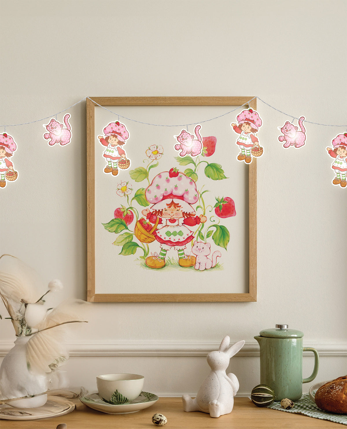 A framed art print featuring a Strawberry Shortcake illustration hangs on a cream wall above a styled tabletop with a white rabbit figurine, a green mug, and other decorative items. A pink and red strawberry-themed string light banner is also visible across the top of the frame.