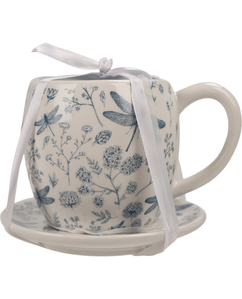 A small ceramic teacup and saucer set in white with a blue botanical and dragonfly pattern, wrapped in a white satin ribbon, on a plain white background.