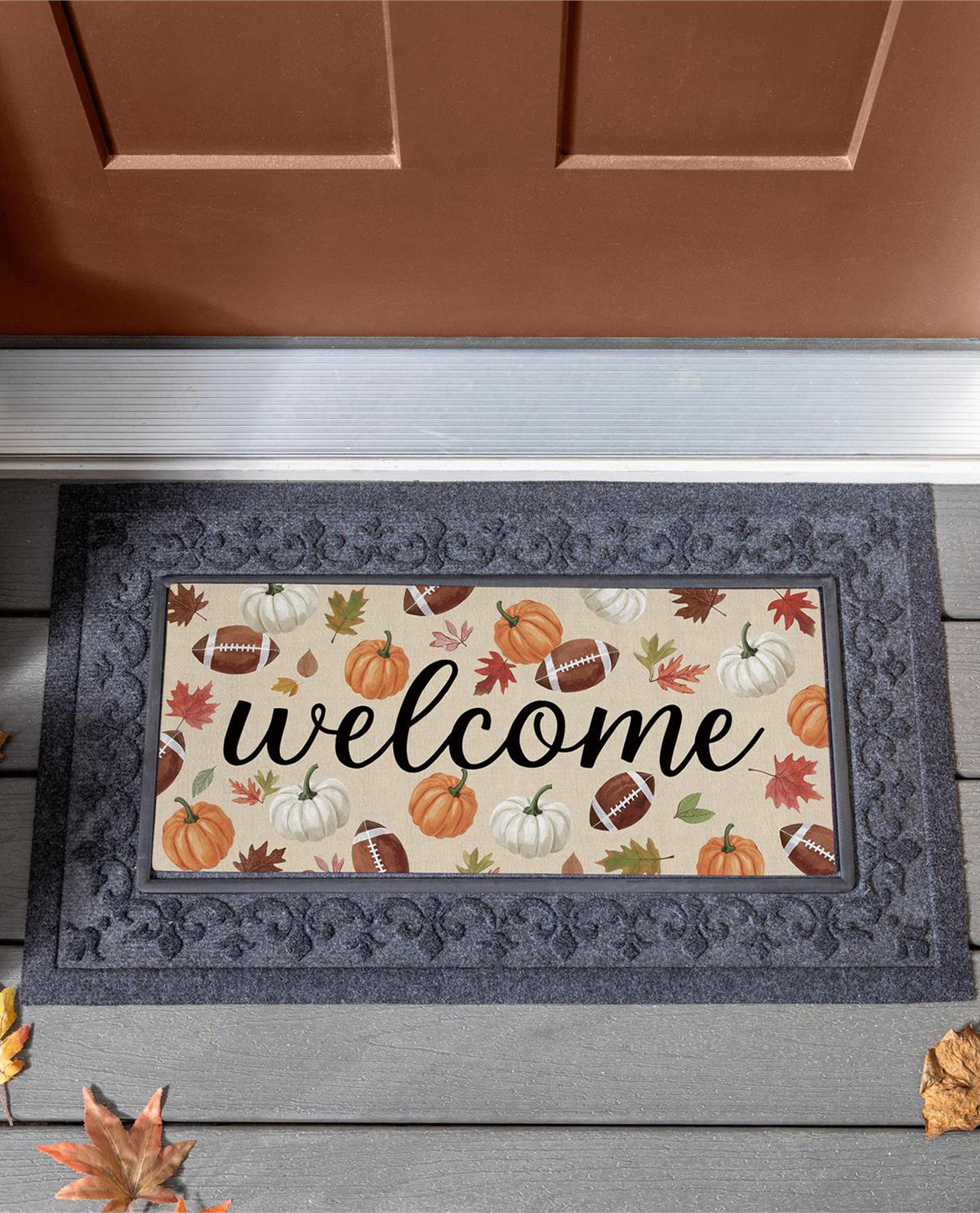 A rectangular doormat with a dark gray ornate border and a cream center featuring the word 'welcome' in black script, surrounded by autumn pumpkins, footballs, leaves, and acorns. The mat is shown on a gray wooden porch in front of a brown door.