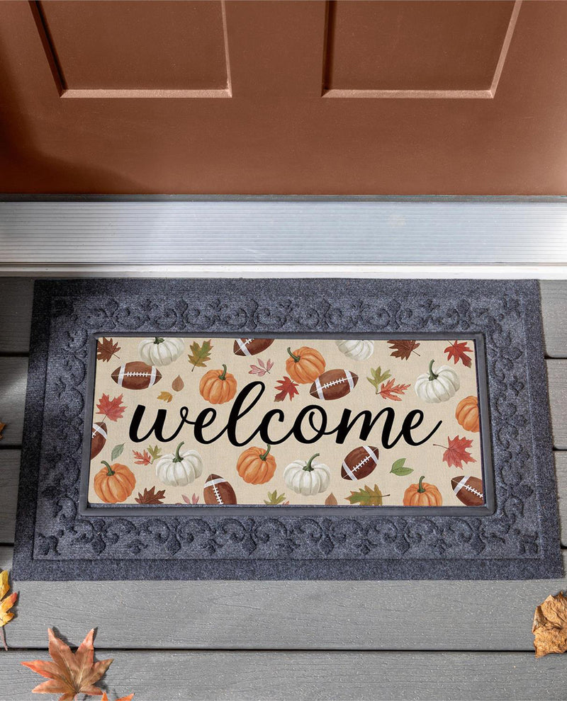 A rectangular doormat with a dark gray ornate border and a cream center featuring the word 'welcome' in black script, surrounded by autumn pumpkins, footballs, leaves, and acorns. The mat is shown on a gray wooden porch in front of a brown door.