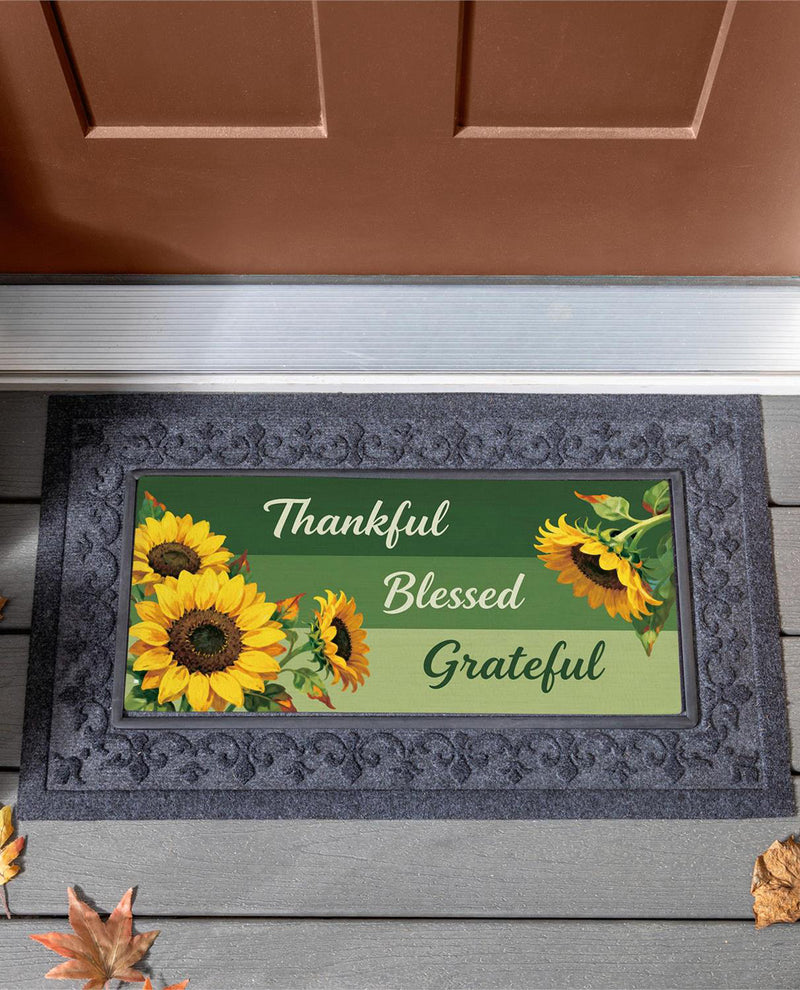 A rectangular doormat with a dark gray ornate border and a printed center featuring yellow sunflowers and the words 'Thankful Blessed Grateful' in white and green text, placed on a gray porch step in front of a brown door.