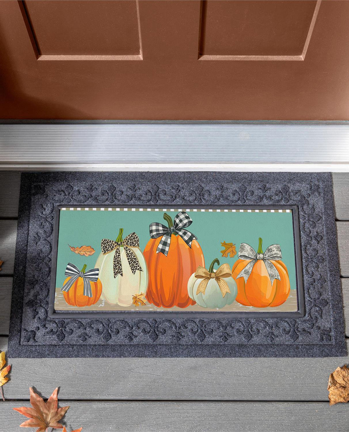 A rectangular doormat with a fall harvest design featuring orange and white pumpkins with bows, set on a dark gray damask border. The mat is shown on a gray wooden porch step in front of a brown door.