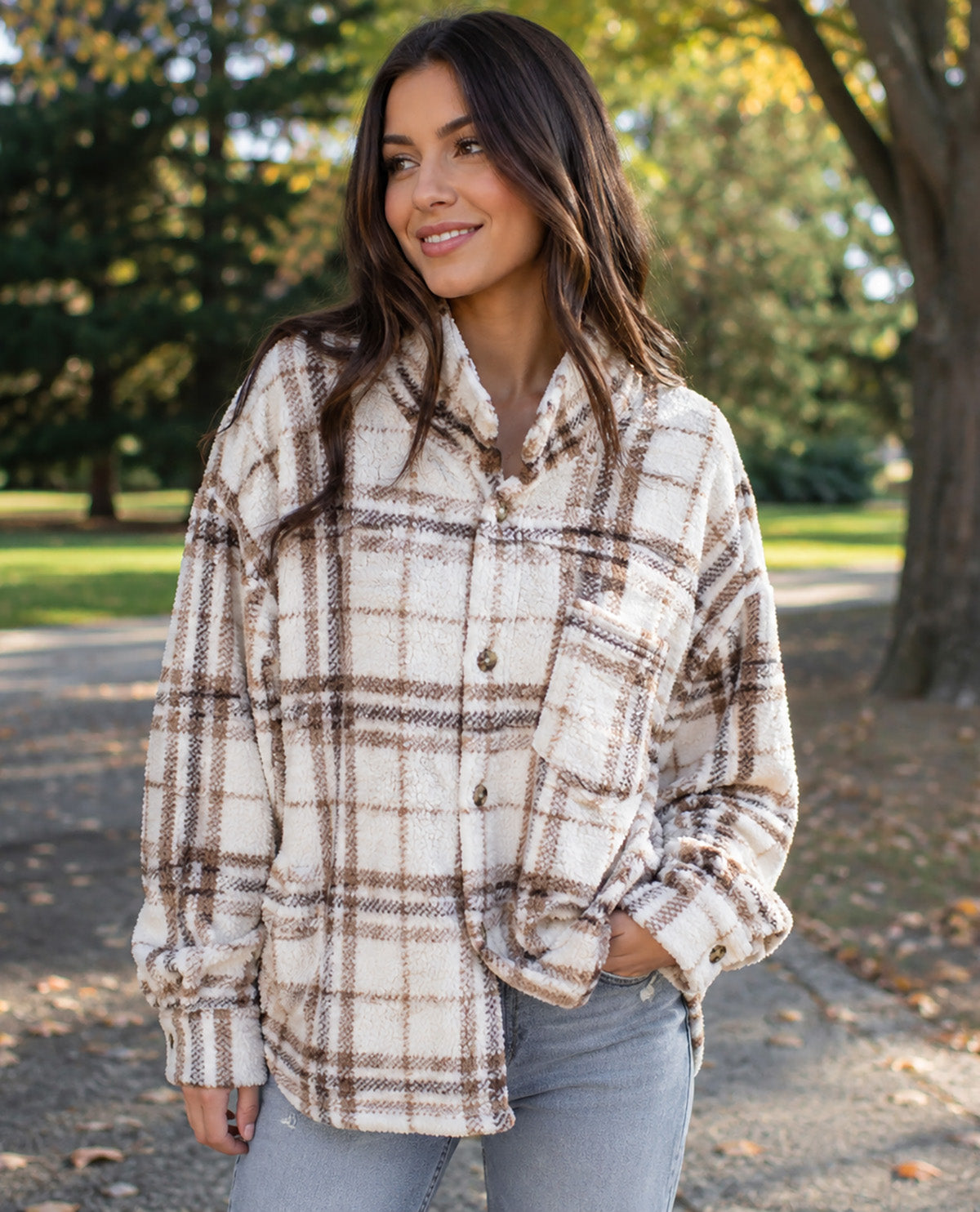 A woman wearing a cream and brown plaid button-up shirt jacket with a soft fleece texture, standing outdoors on a path with trees in the background.
