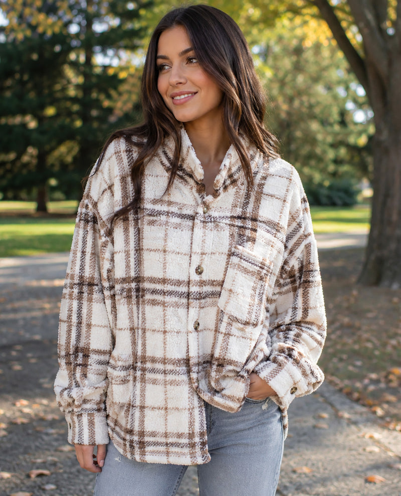 A woman wearing a cream and brown plaid button-up shirt jacket with a soft fleece texture, standing outdoors on a path with trees in the background.