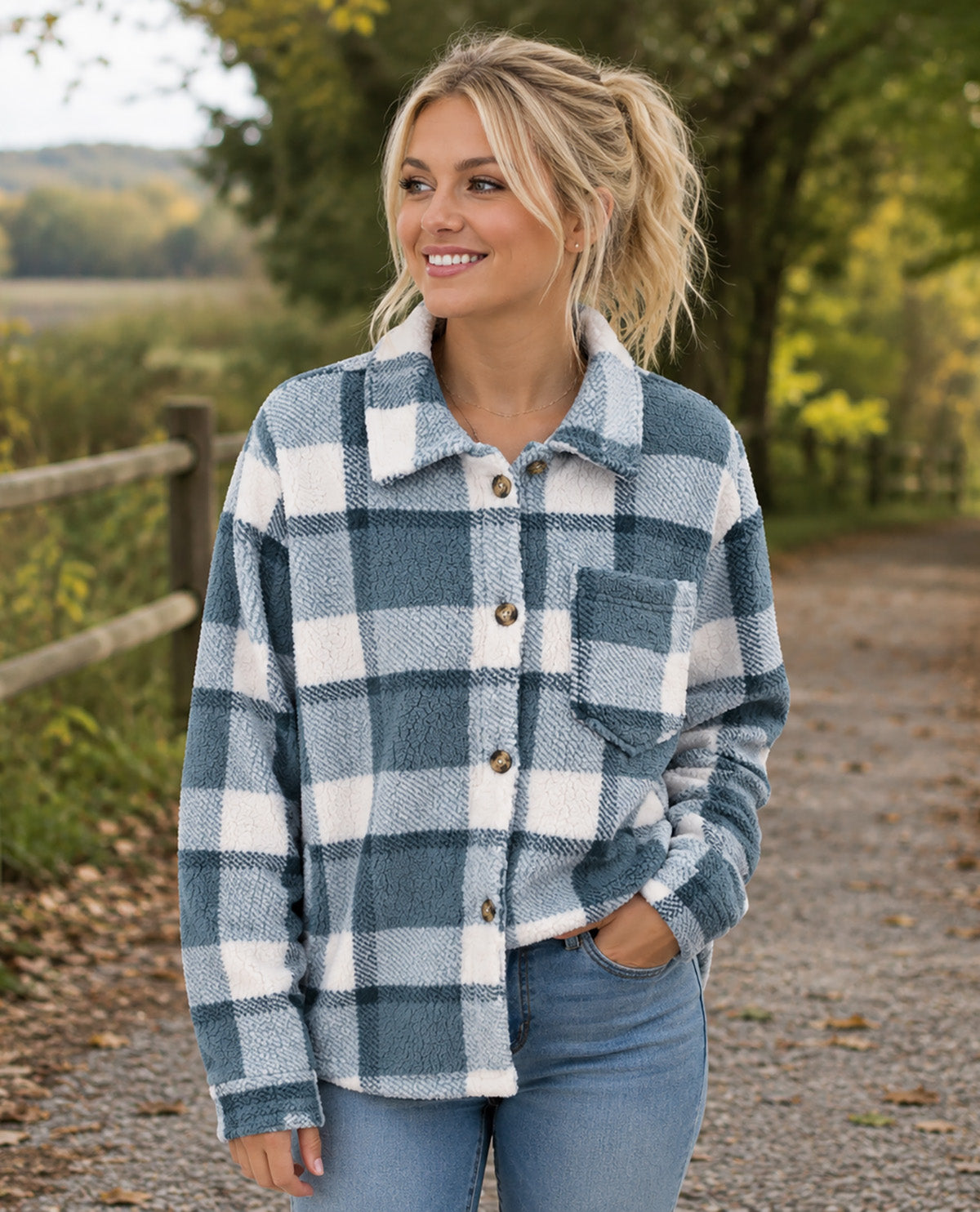 A woman wearing a blue and white plaid flannel button-up shirt jacket with blue jeans, standing on a gravel path outdoors with trees and a fence in the background.