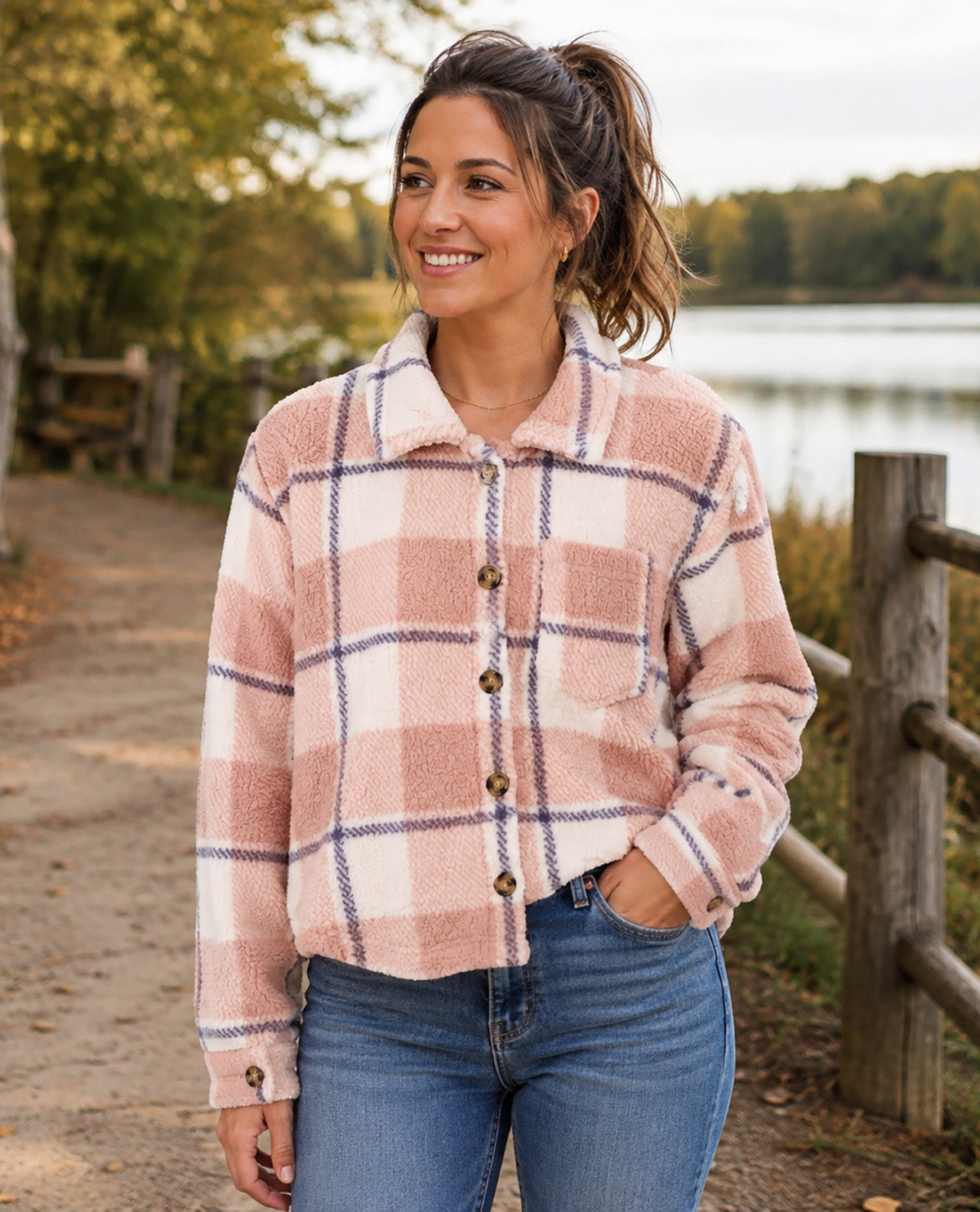 A woman wearing a pink and white plaid fleece button-up shirt with blue jeans, standing on a path near a lake with a wooden fence in the background.