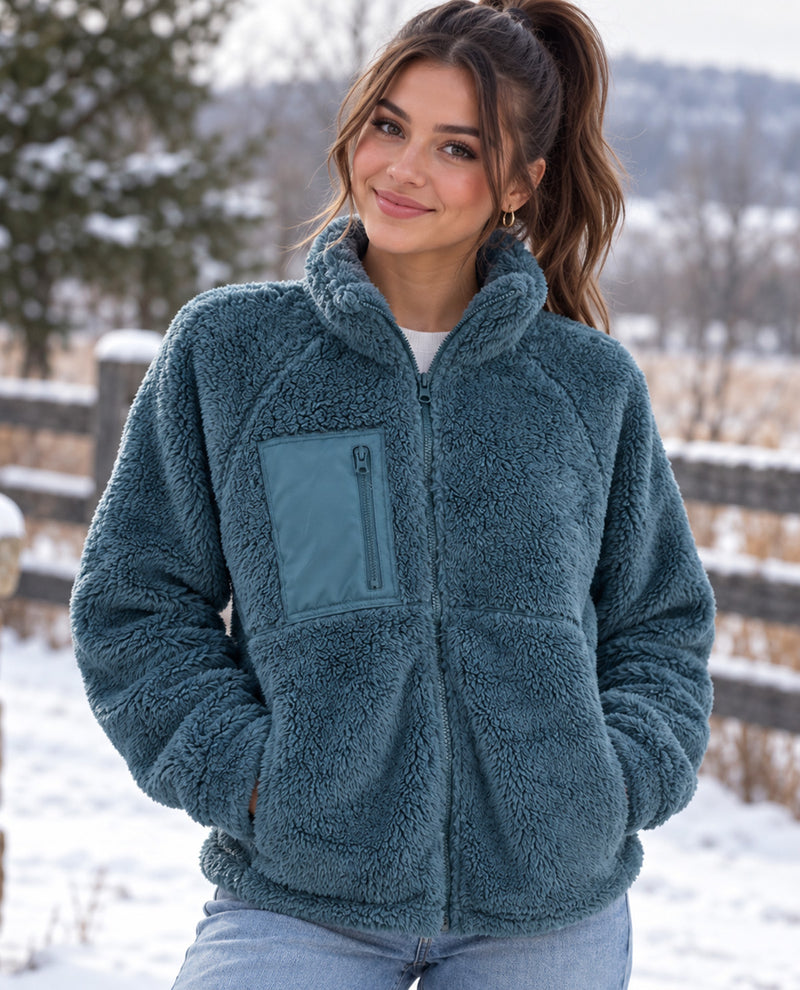 A woman wearing a teal blue sherpa fleece zip-up jacket with a chest pocket, standing outdoors in a snowy landscape.