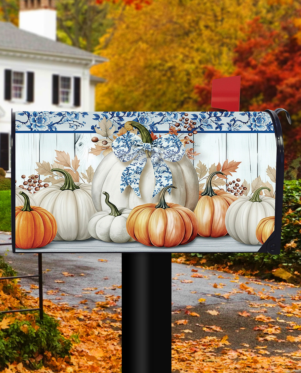 A decorative mailbox cover featuring a fall harvest design with white and orange pumpkins, blue floral trim, and a blue bow, shown installed on a black mailbox post in front of a house and autumn trees.