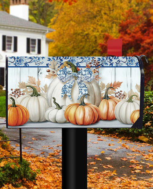 A decorative mailbox cover featuring a fall harvest design with white and orange pumpkins, blue floral trim, and a blue bow, shown installed on a black mailbox post in front of a house and autumn trees.