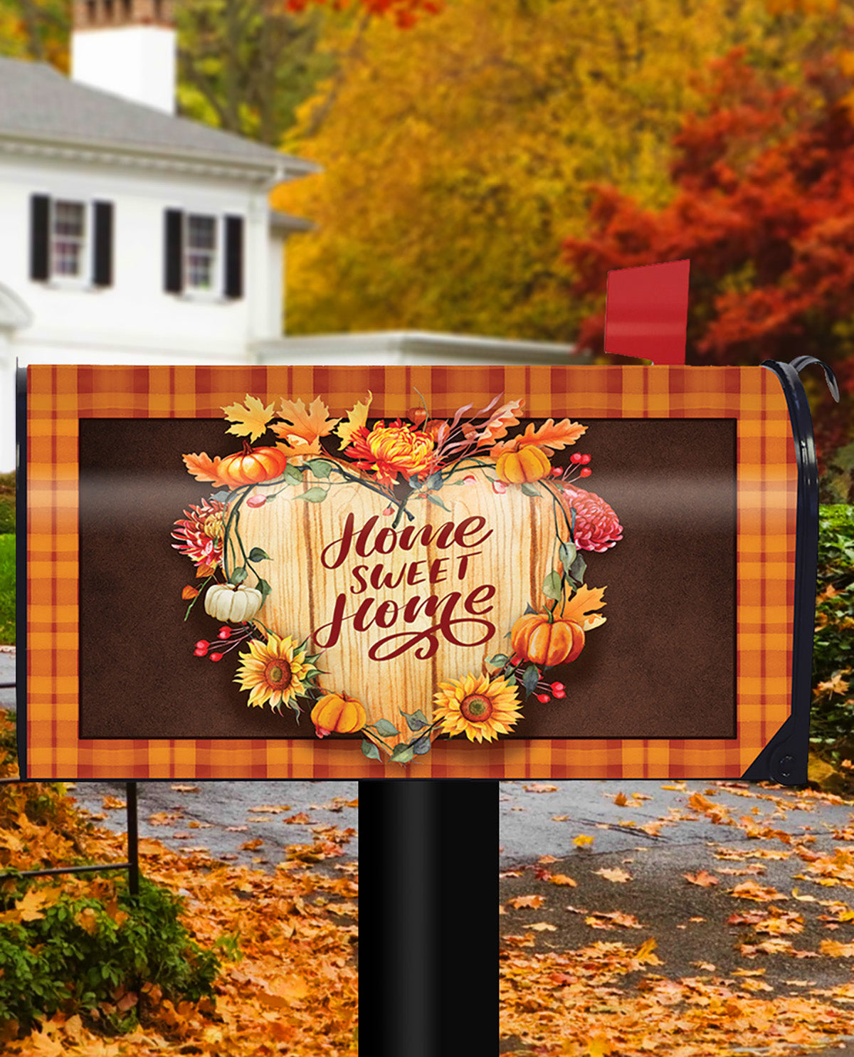 A decorative mailbox cover with a fall harvest design, featuring the phrase 'Home Sweet Home' in cursive over a wood-grain pumpkin motif, surrounded by pumpkins, sunflowers, and autumn leaves. The cover has an orange plaid border and is shown mounted on a black mailbox post in a residential outdoor setting with a house and colorful autumn trees in the background.