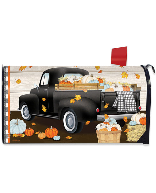 A decorative mailbox cover featuring a vintage black pickup truck loaded with pumpkins and gourds, with scattered autumn leaves, pumpkins, and a basket of produce on a rustic wood-plank background. A red flag is visible on the right side.