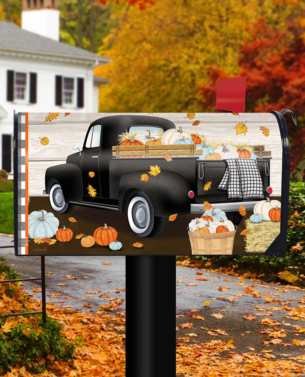 A decorative mailbox cover featuring a vintage black pickup truck loaded with pumpkins and fall leaves, shown installed on a mailbox in an autumn yard with a house and trees in the background.