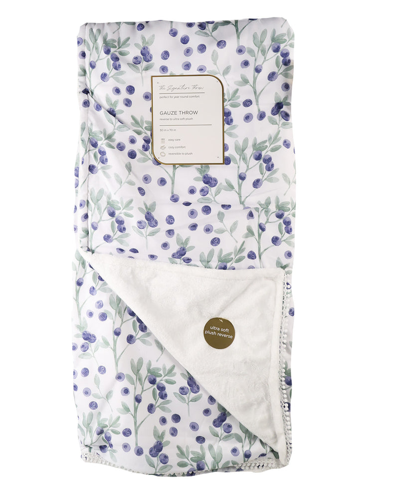 A folded baby blanket with a white background and a blueberry and green leaf pattern, featuring a cream-colored plush reverse side and a paper brand label.