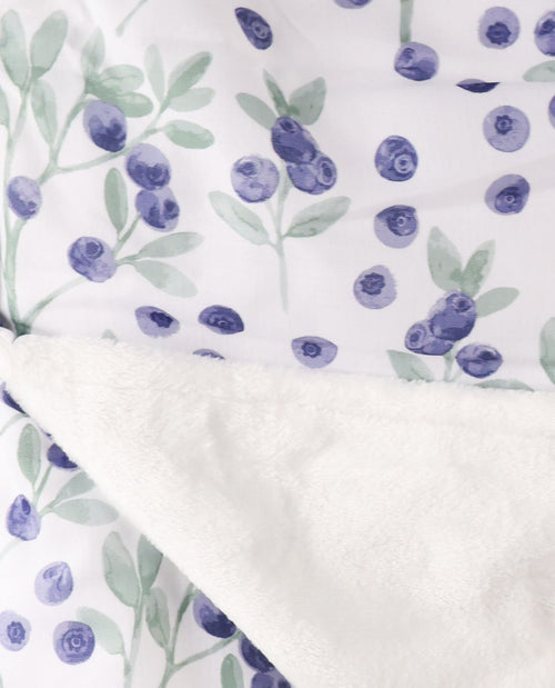 Close-up of a soft textile product with a white base printed with watercolor-style blueberries in purple and green, plus a folded white sherpa-like fleece layer.