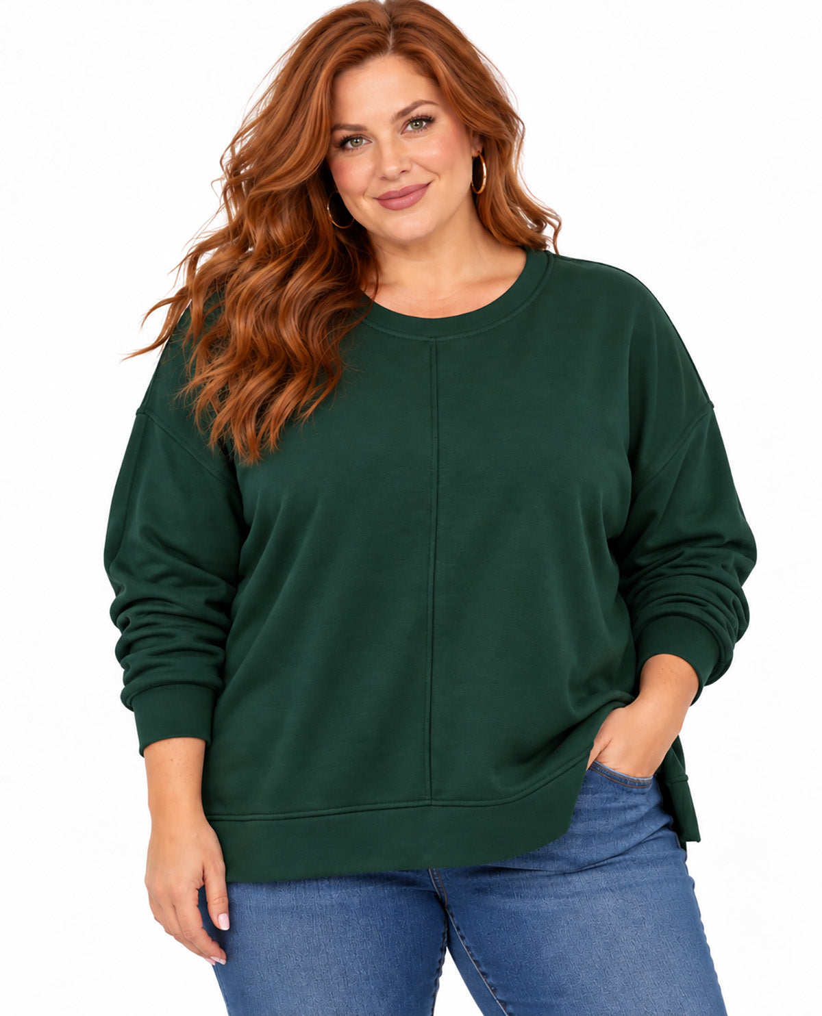 A woman with long wavy red hair wearing a dark forest green crewneck sweatshirt and blue jeans, standing against a plain white background.
